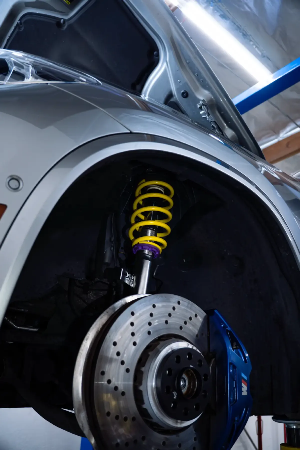 Yellow coilover spring and blue brake caliper behind a drilled rotor, seen in a car's front wheel well.