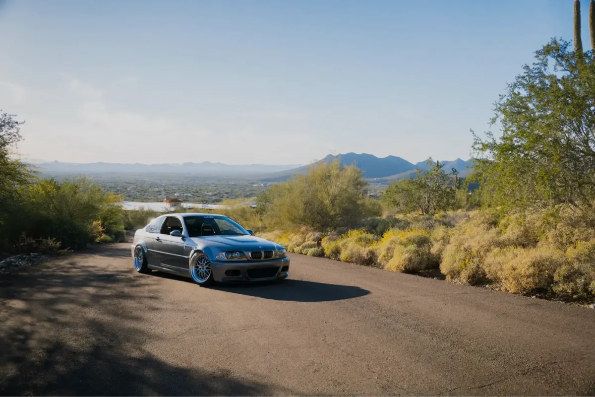 Grey BMW M3 (E46) driving a winding desert mountain road at golden hour.