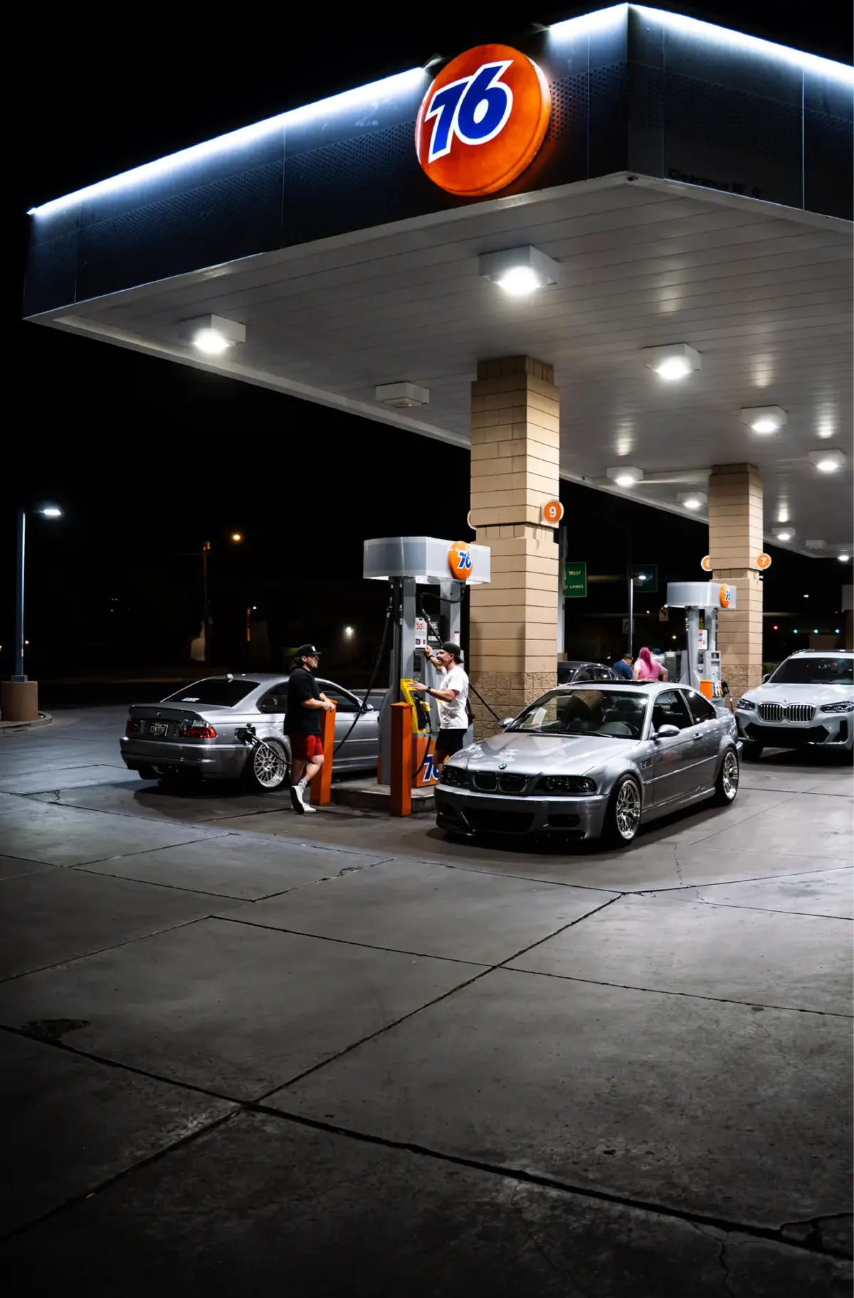 Two silver E46 M3 coupes parked under a brightly lit gas station canopy at night