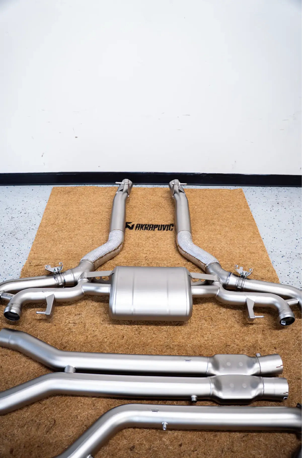 A complete Akrapovič exhaust system laid out on a mat on the shop floor, with muffler, heat-shielded pipes, hangers and tips