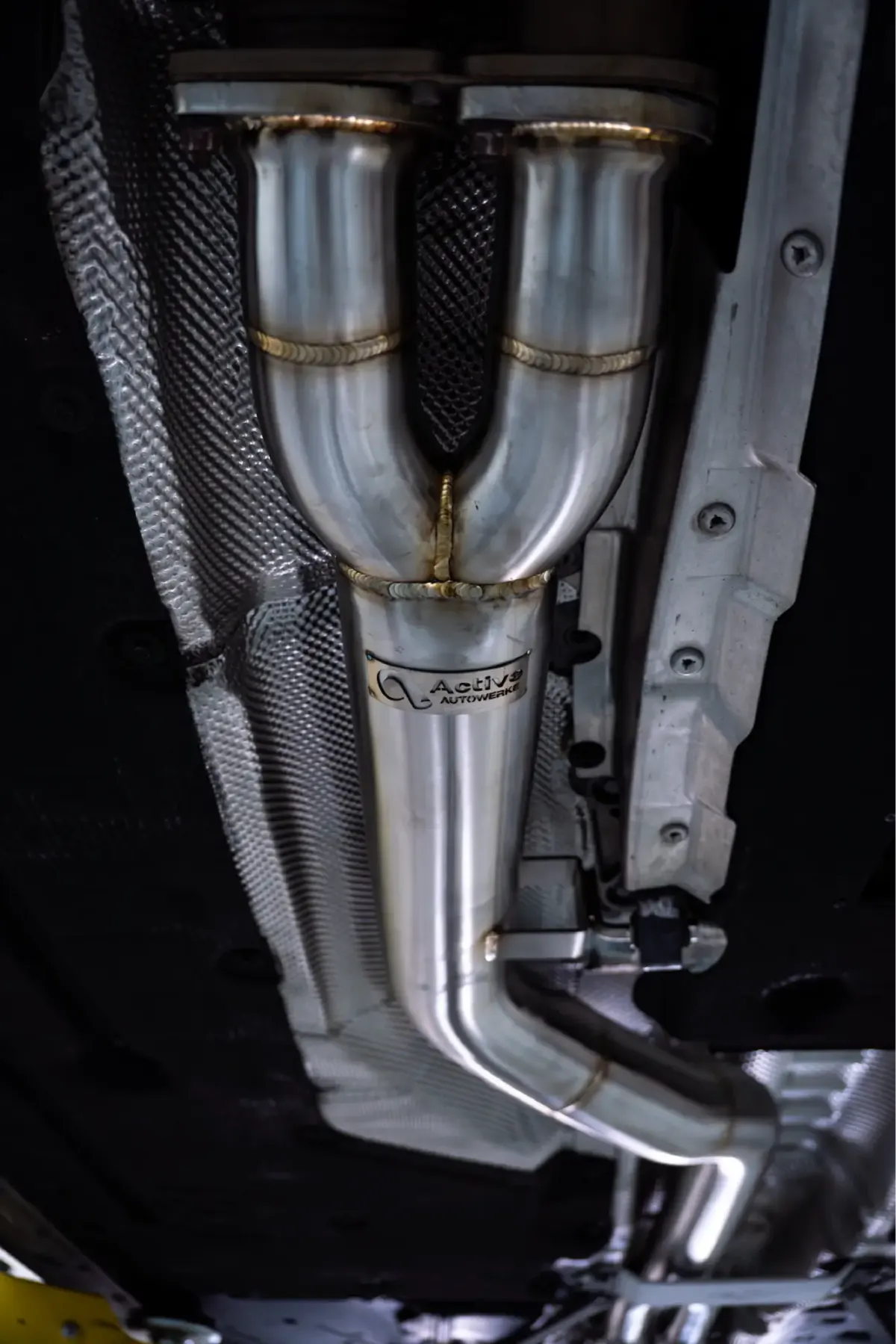 Stainless Active Autowerke exhaust section with twin pipes merging into one, seen from underneath a car next to the heat shielding