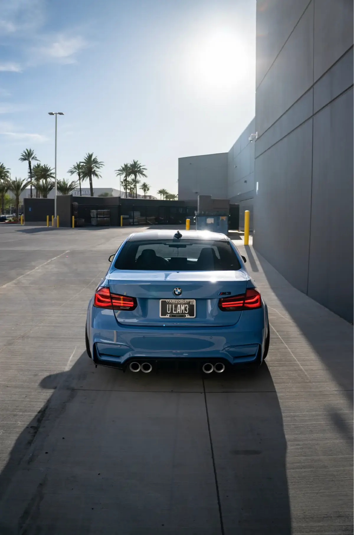 Rear view of a blue BMW M3 (F80) sedan between a grey building and palm trees.