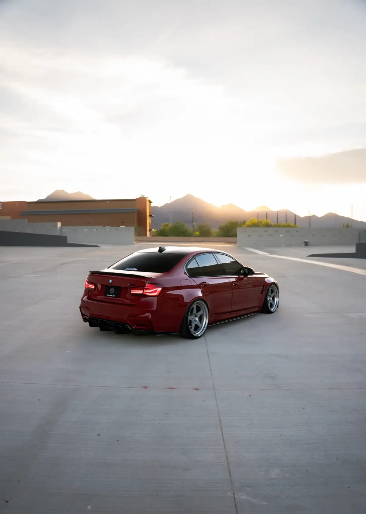 Maroon BMW M3 (F80) sedan, rear three-quarter view, on a rooftop parking deck at sunset with mountains on the horizon.