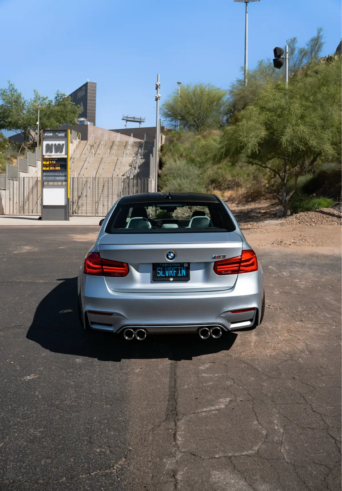 Rear view of a silver BMW M3 (F80) on a desert road with a stadium light tower behind it.