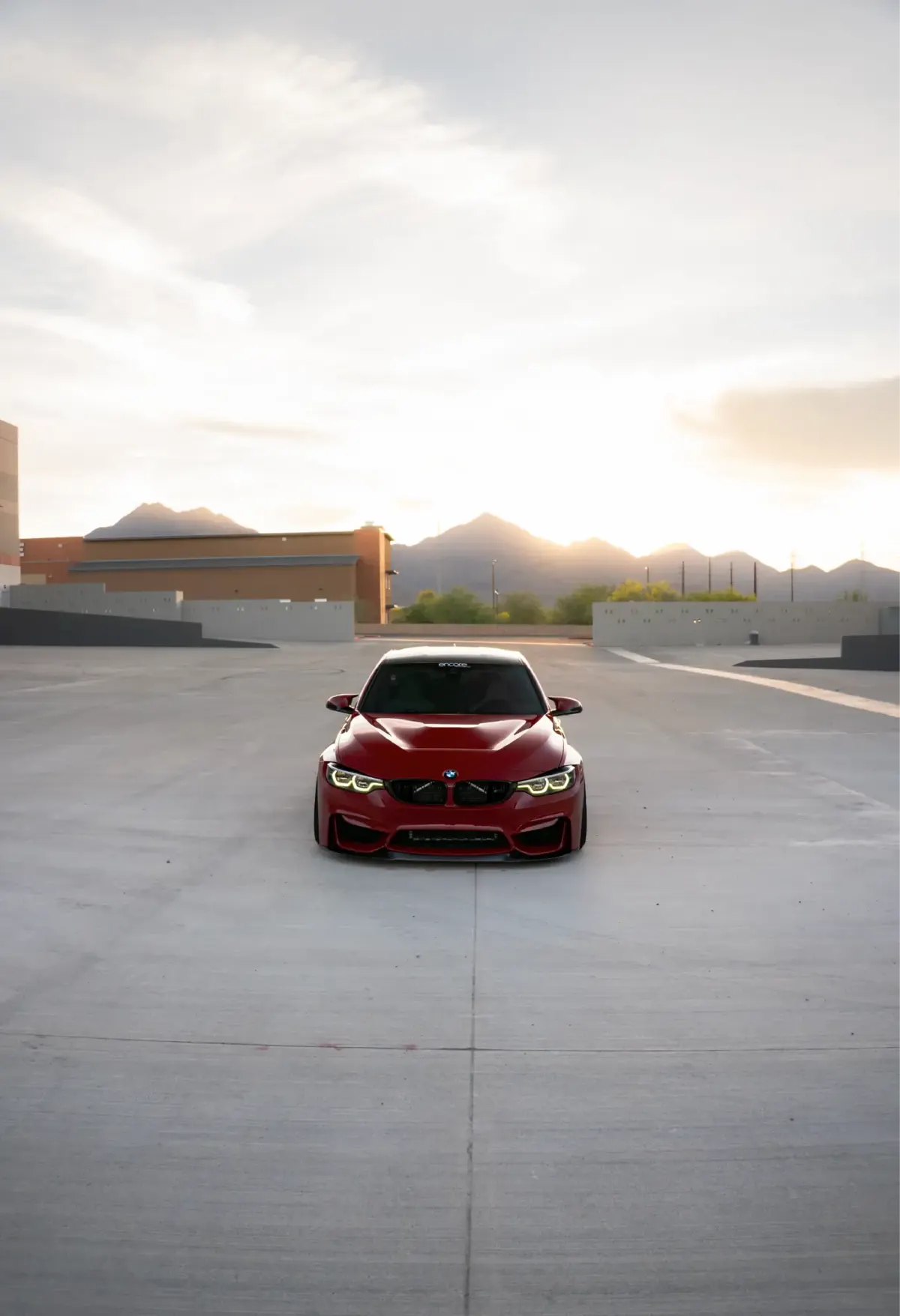 Red BMW M3/M4 (F8x) facing the camera on an empty rooftop deck at sunset, mountains behind.