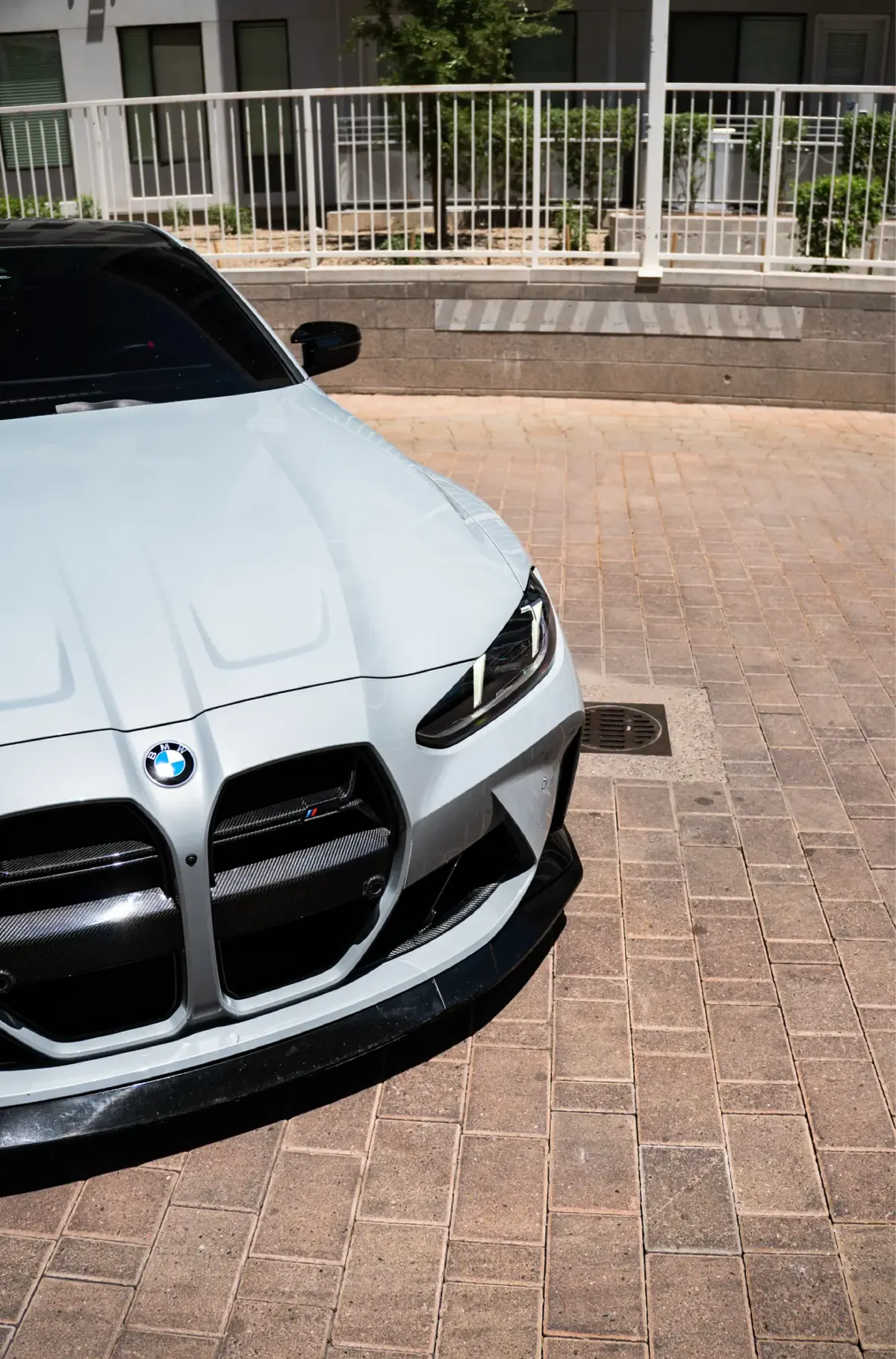 Close-up of a white BMW M4 (G82) front grille with dark slats and a carbon-look front lip, on a brick plaza