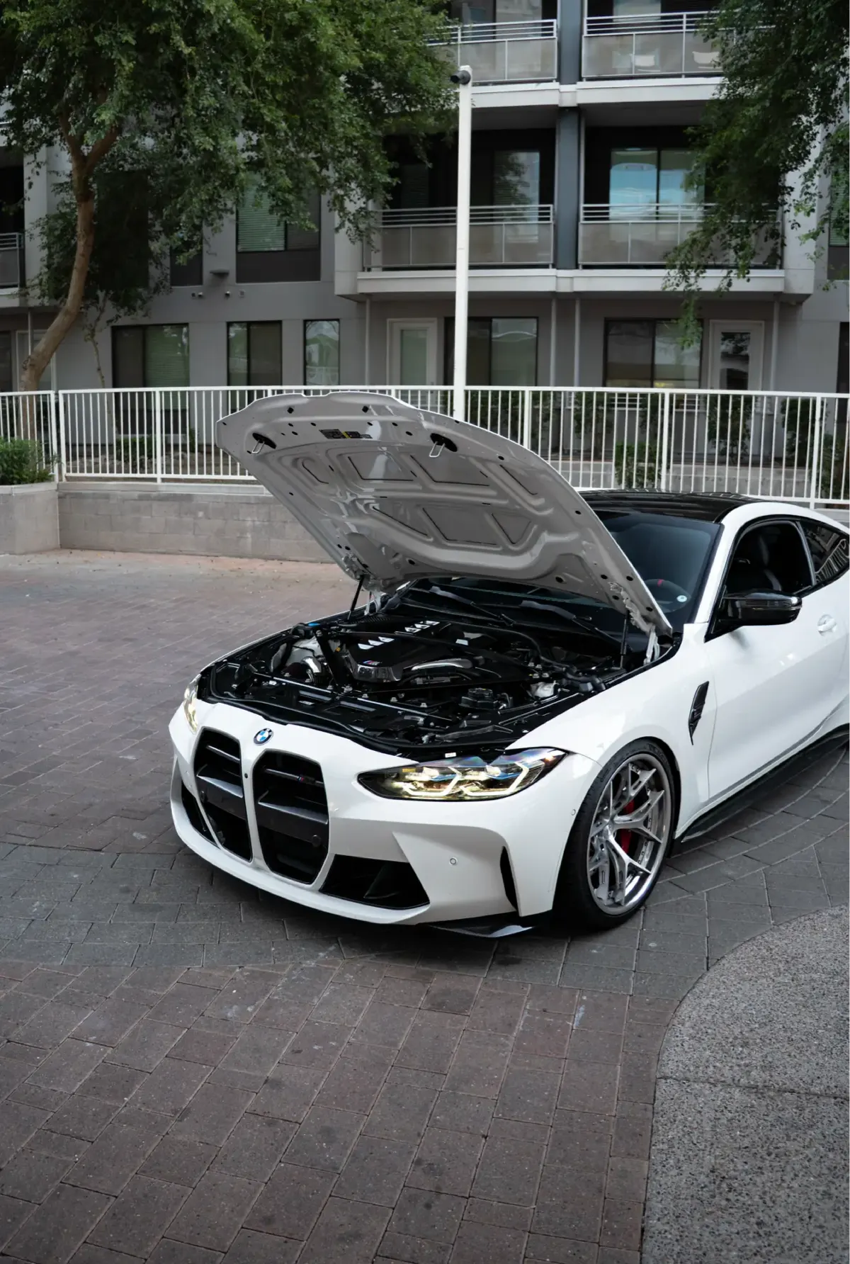 White BMW M4 (G82), front three-quarter view, hood raised outside an apartment building