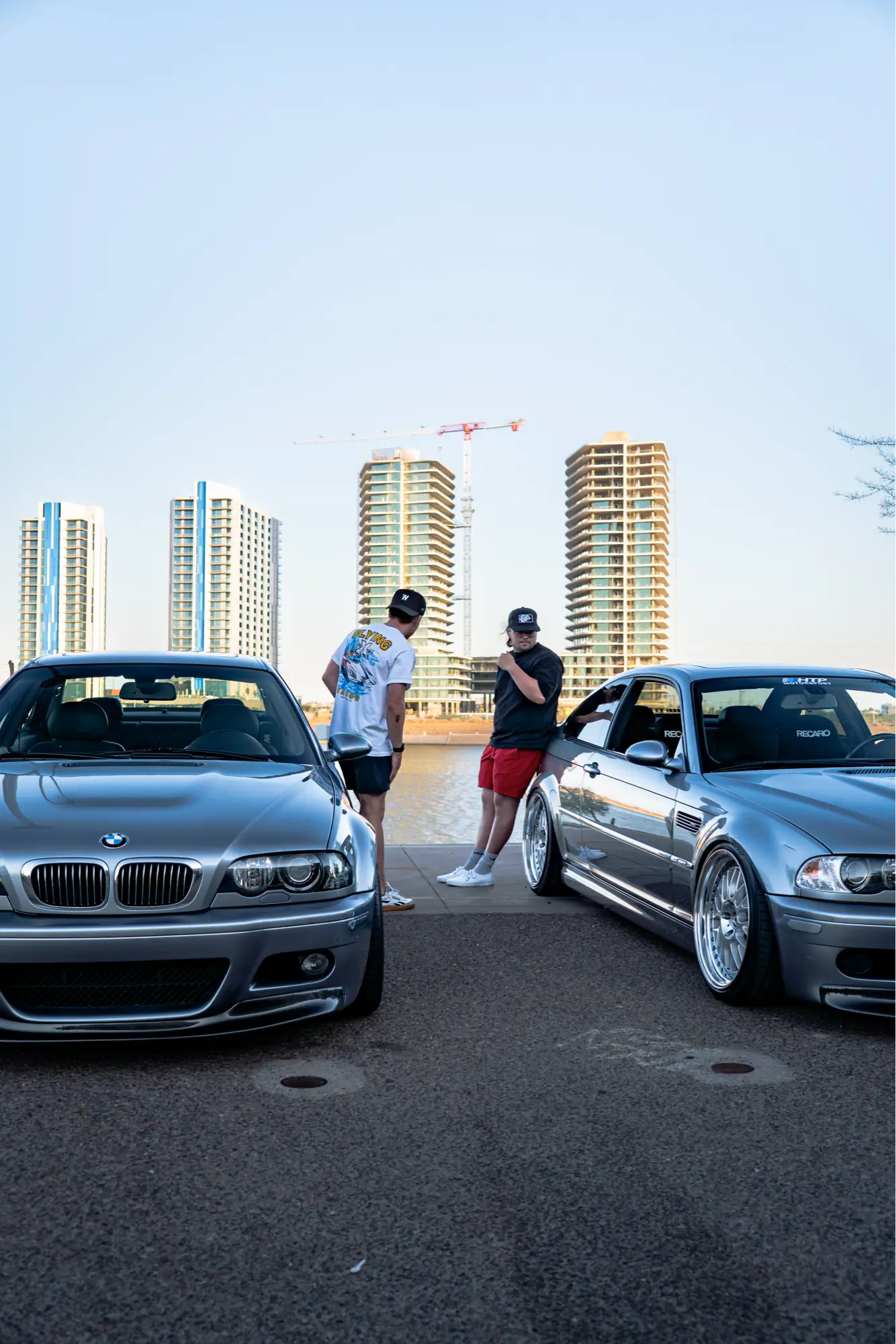 Close shot of a pair of silver BMW E46 M3s parked along a riverfront with the city skyline behind them.