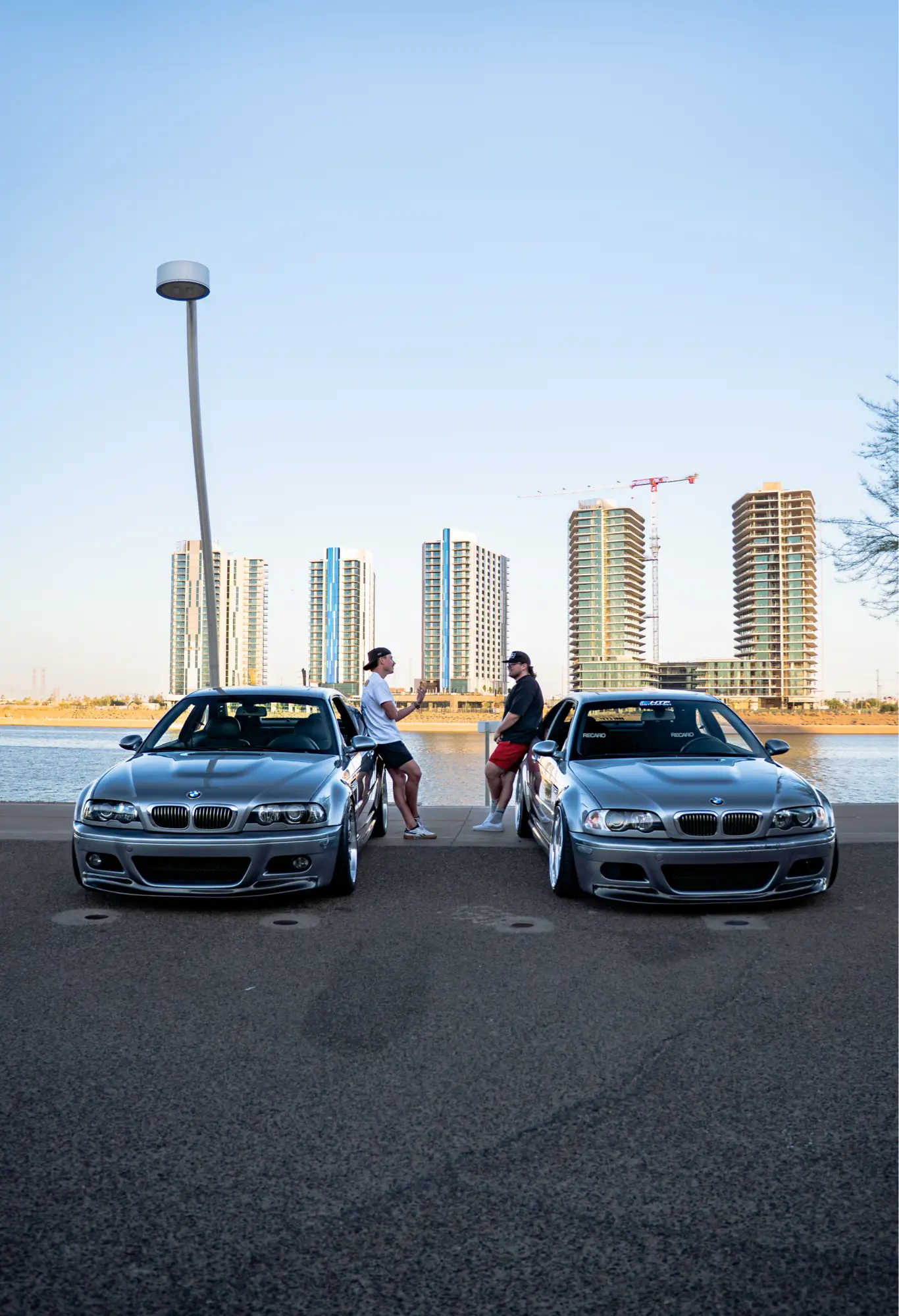 A pair of silver BMW E46 M3s parked along a riverfront, city skyline visible in the distance.