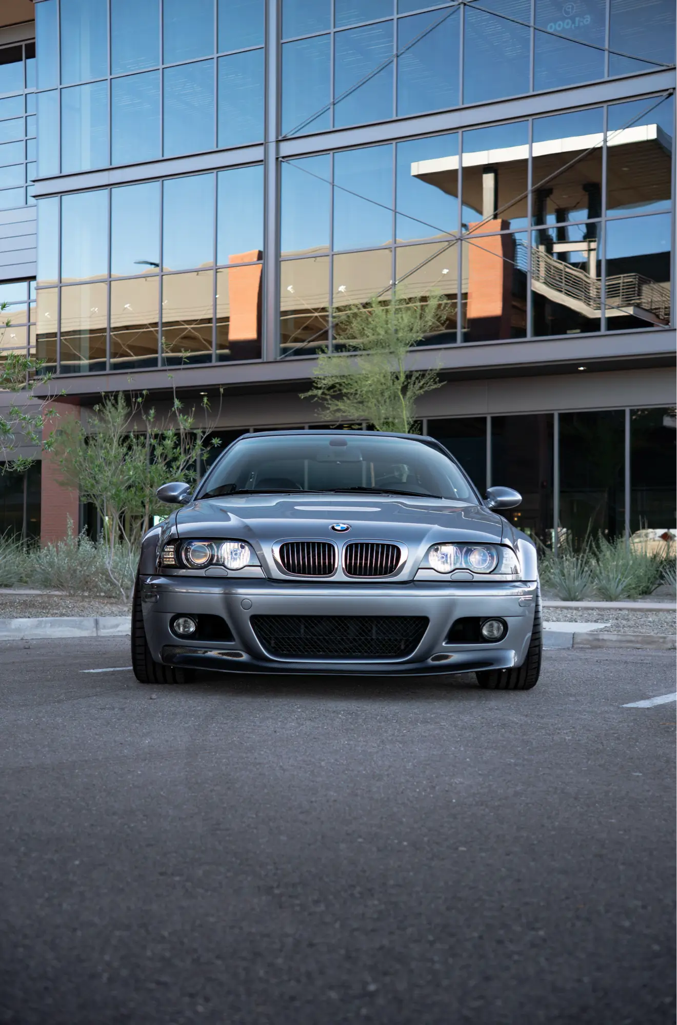 Straight-on front view of a silver BMW E46 M3 parked outside an office building.