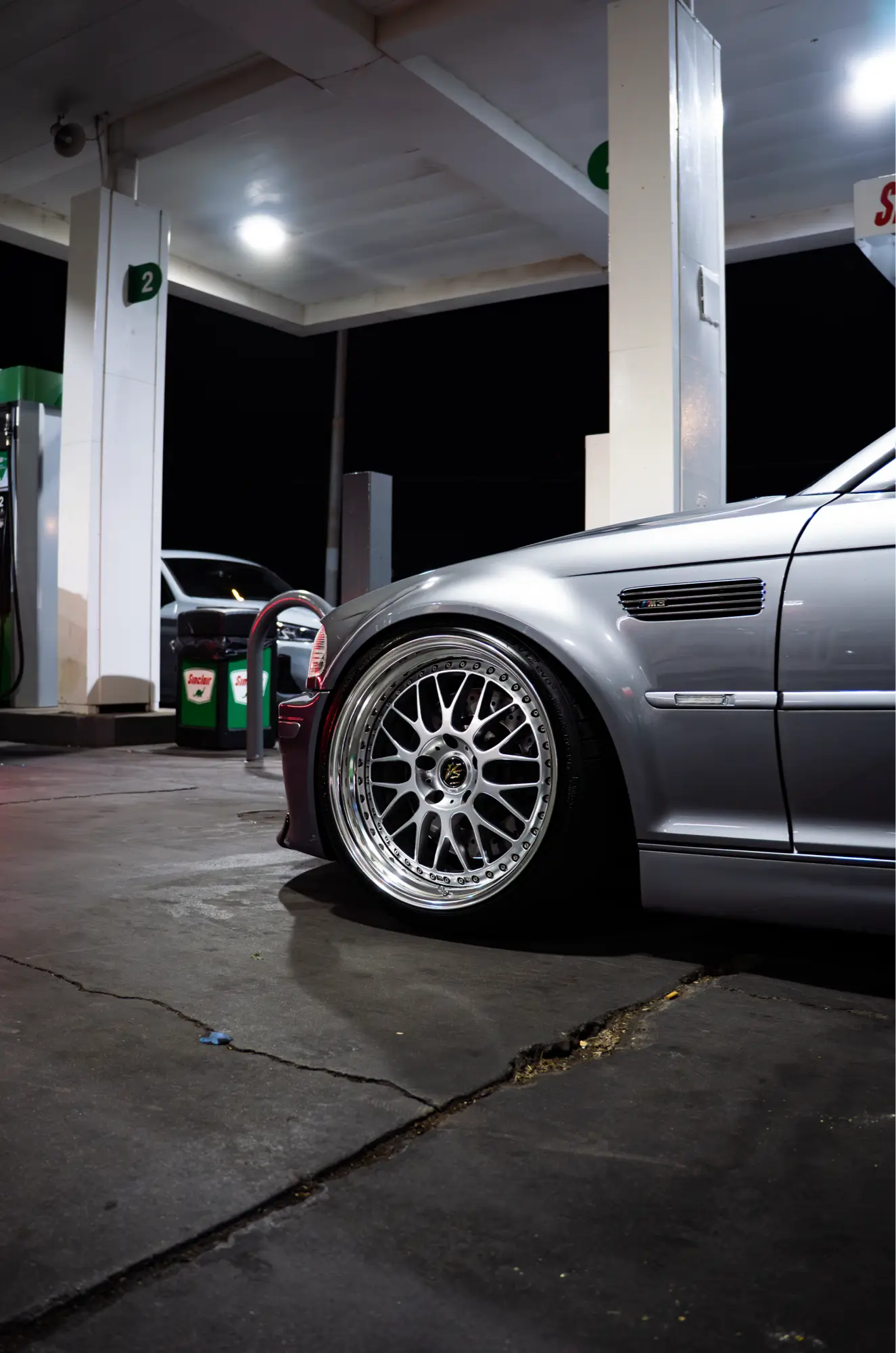 Close-up of a silver BMW E46 M3's wheel at a gas station at night.