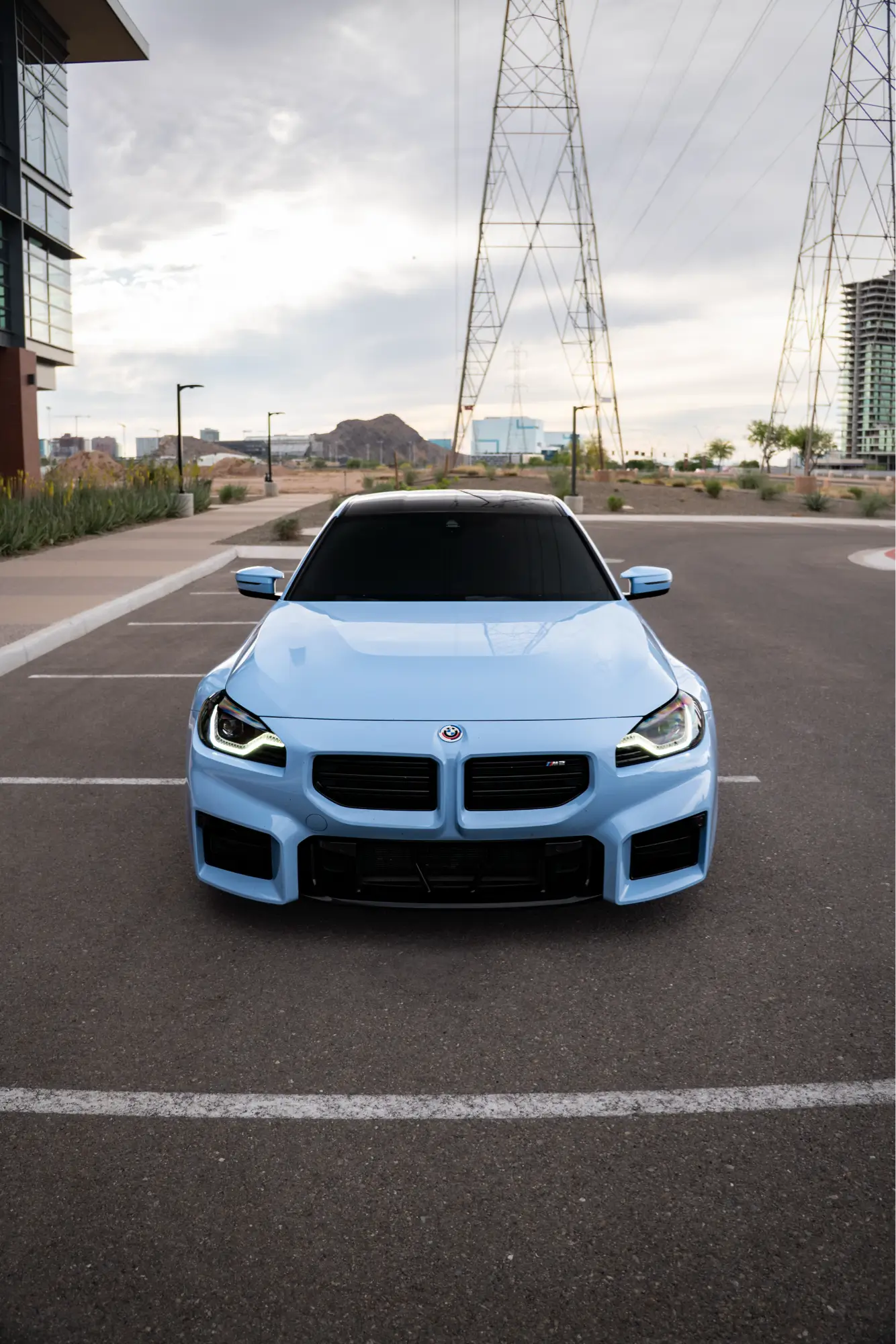 Straight-on front view of a blue BMW M2 (G87) with transmission towers behind it.