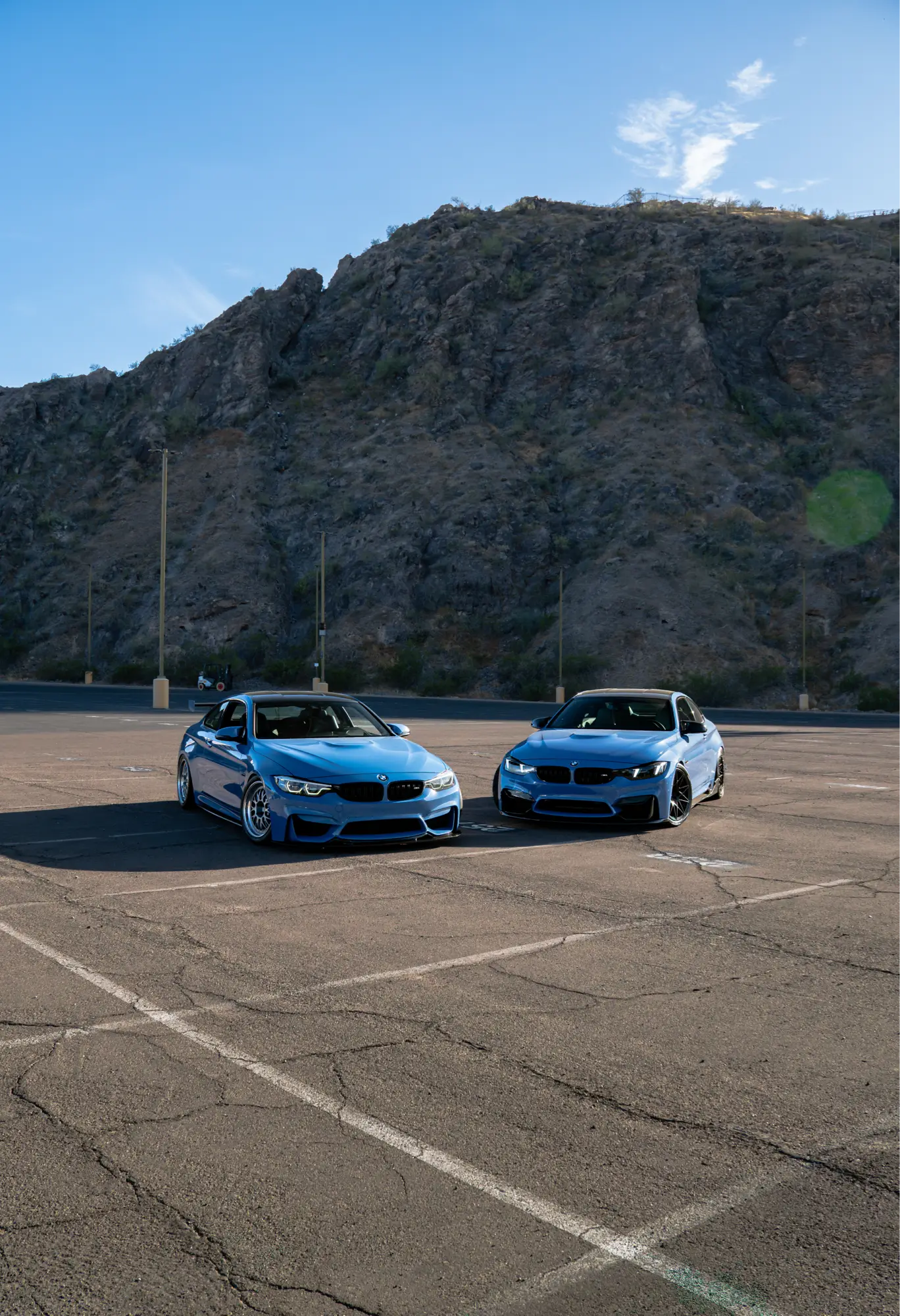 A pair of blue BMW M3/M4 (F80/F82) models parked together in a lot with mountains behind them.