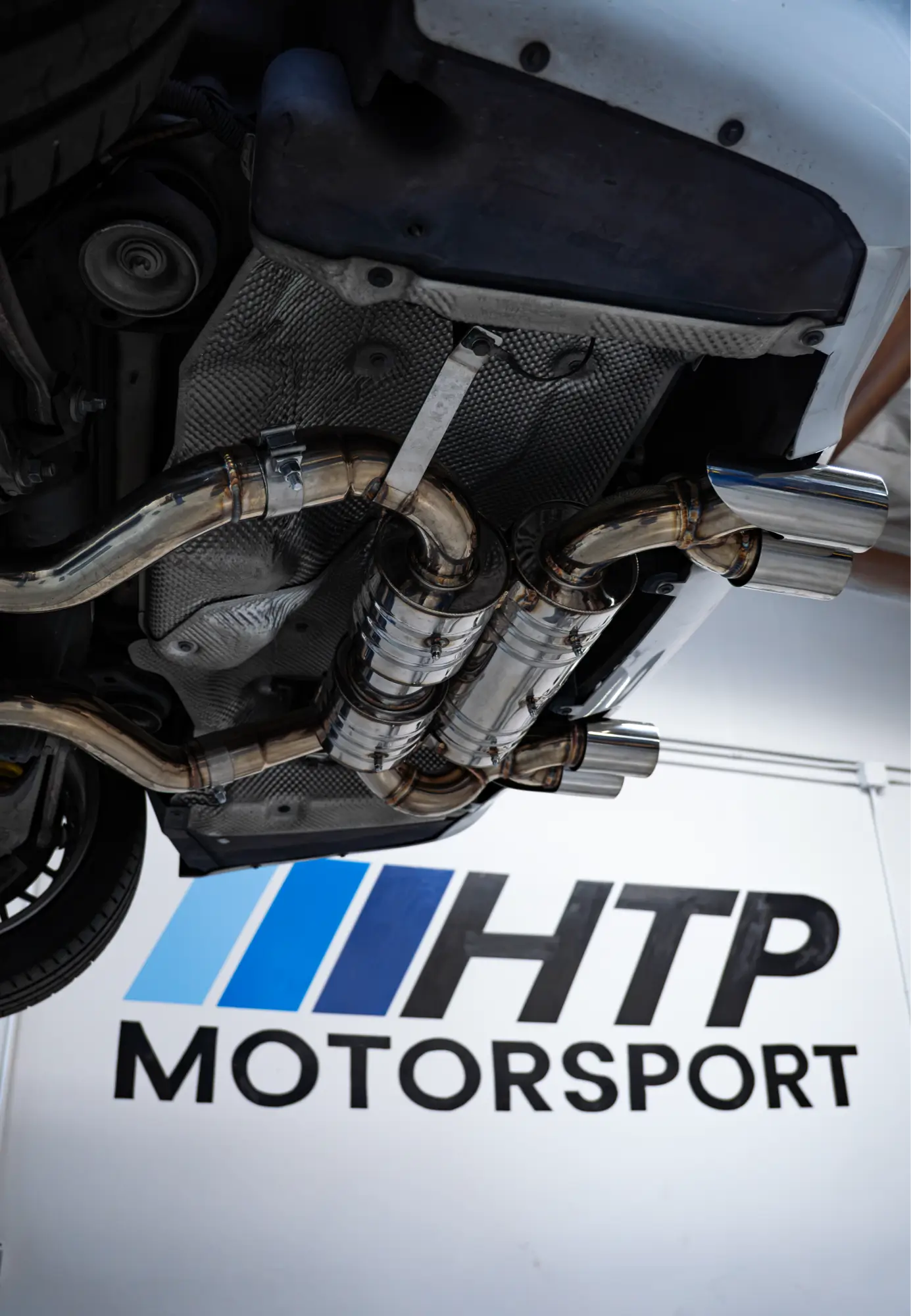 Exhaust installation underway underneath a white car at the HTP Motorsport shop.