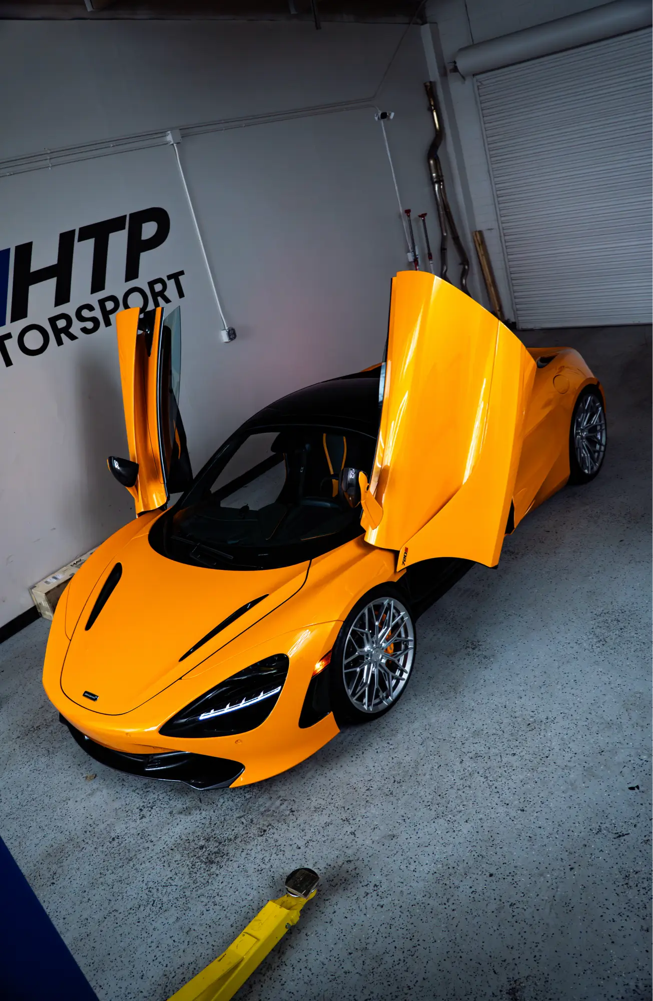 Orange McLaren 720S with its dihedral doors raised, shown front three-quarter beneath an overhead door.