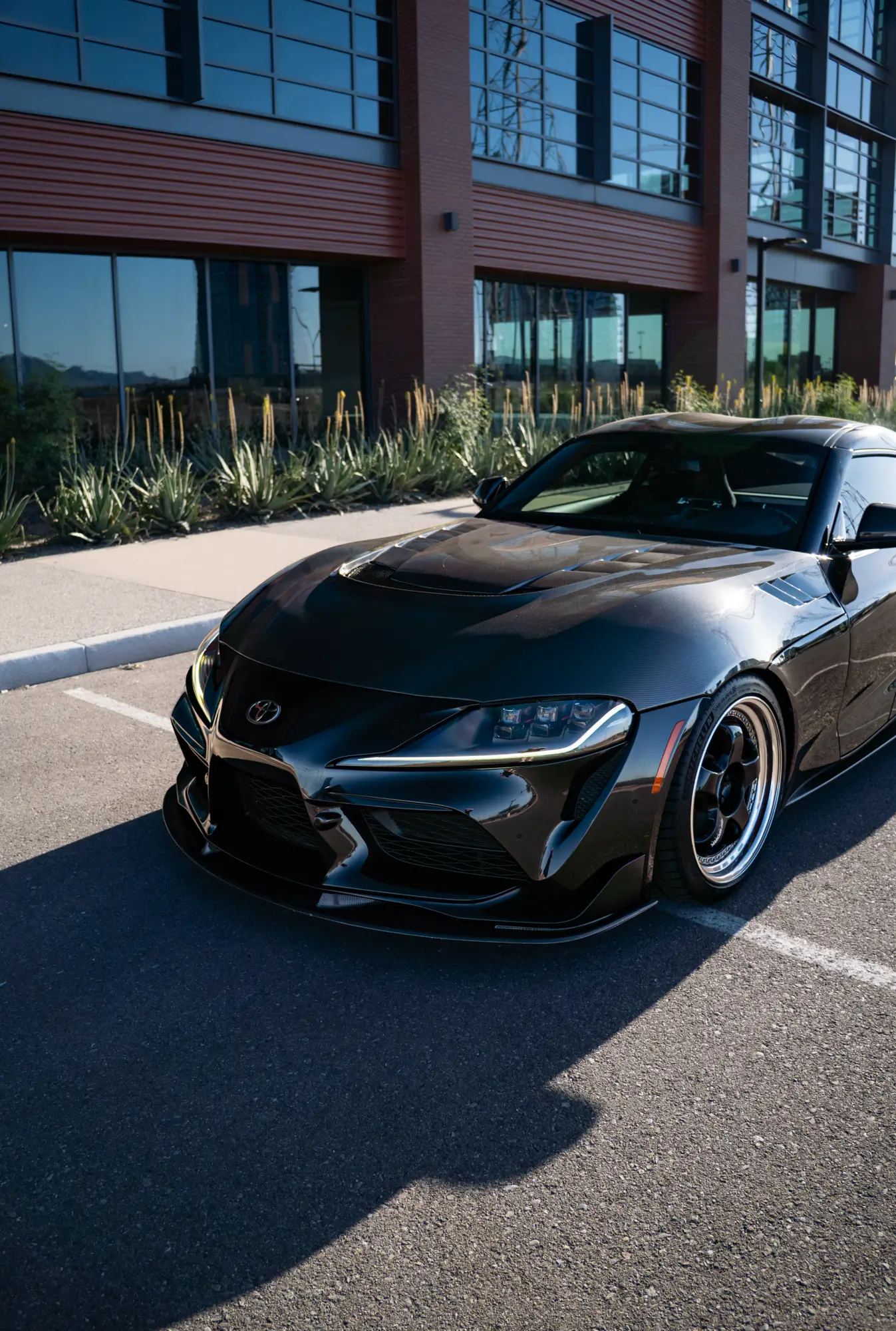Black Toyota Supra (MK5) shown front three-quarter in an office plaza.